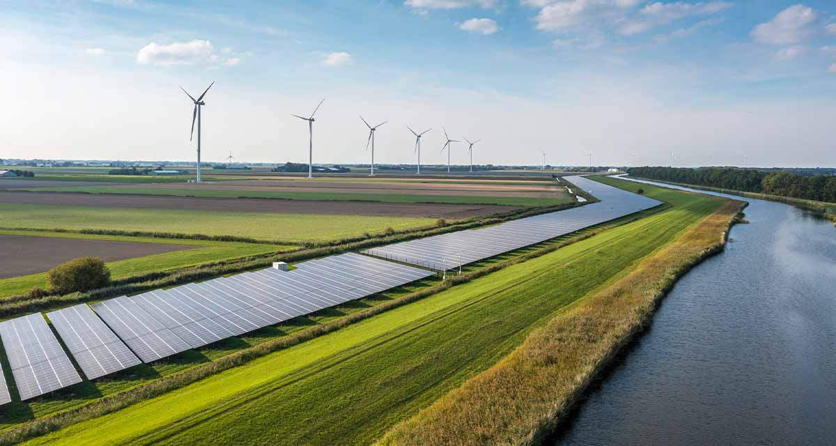 Paesaggio di campagna con campo fotovoltaico accanto a un fiume e pale eoliche: un'alleanza vincente per la sostenibilità energetica